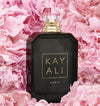 A black perfume bottle with a faceted clear cap and gold lettering reading 'KAYALI ELIXIR 11', surrounded by pink flower petals.