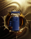A blue cylindrical perfume bottle with gold detailing and a tassel, labeled 'HISTORIC OLIMPA BY A'NAN', centered within a metallic gold liquid splash on a dark reflective background.