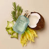 A bottle of KAYALI Salting the Rain Coconut & Rosemary perfume, styled in a flat-lay with a halved coconut, starfruit, yellow flowers, and rosemary sprigs on a warm beige background.