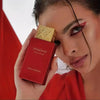 A close-up of a model holding a red Swiss Arabian Shaghaf Old Arabian perfume bottle with a gold cap, set against a red and white background.