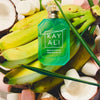 A green glass perfume bottle labeled 'KAYALI' and 'MASH IN A BOTTLE SWEET BANANA 197' placed on top of fresh bananas, coconut halves, and green leaves.