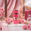 A pink perfume bottle with gold trim and a decorative ribbon label stands on a white marble surface, surrounded by pink roses and draped pink fabric in a styled indoor setting.