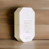 A white octagonal box of Kay Ali Sea Yu White Flower Lip Eau de Parfum, standing upright on a light wood surface against a darker wood background.