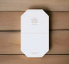 A white octagonal folded card or envelope with small gold text, photographed from above on a light brown wood plank surface. The top half has a list of ingredients or notes, and the bottom half is blank.
