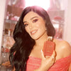 A woman with long, wavy black hair holds a terracotta-colored perfume bottle labeled 'KAYALI'. She is wearing a sparkling coral-pink off-the-shoulder top, with a softly blurred room in the background.