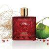 Red Versace perfume bottle with gold cap, green fruit, and decorative elements on a white background